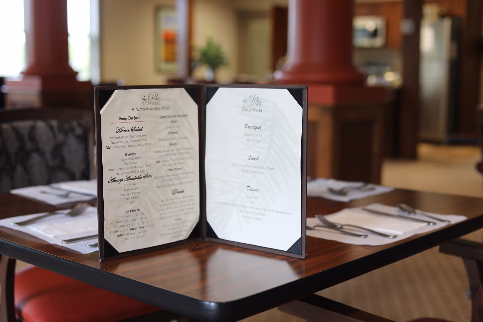 A dining table set with silverware and napkins, featuring an open menu from The Pines Senior Living placed in the center. The background shows a warm, softly lit interior with wooden columns and furniture.