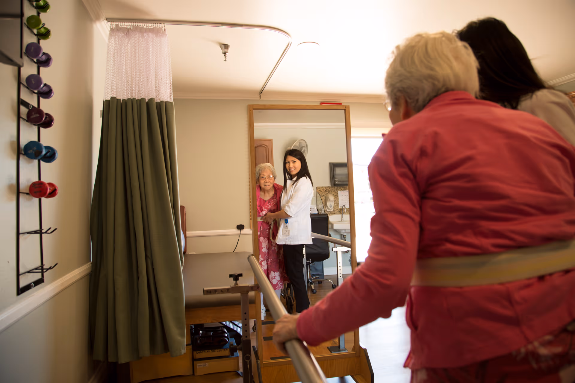 An elderly woman in a pink jacket uses parallel bars for walking assistance in a therapy room. A healthcare professional in a white coat stands with another elderly woman in a pink dress, both visible in a large mirror on the wall. The room has a green privacy curtain and various therapy equipment.