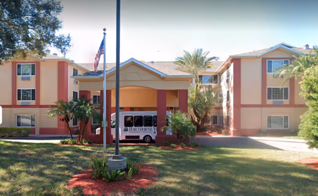 Exterior view of Hawthorne Estates of Brandon, a three-story residential building with beige and red accents. The entrance has a covered driveway with a white shuttle van parked underneath. There are palm trees and landscaped greenery around the building, and an American flag is flying on a flagpole near the entrance.
