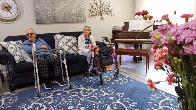 Two elderly individuals sitting on a dark blue couch in a living room. One person is wearing sunglasses and has a walker in front of them, while the other person has a rollator walker nearby. The room features a blue patterned rug, a piano with sheet music and teddy bears on top, a wall clock, a decorative wall piece, and a bouquet of pink and purple flowers in the foreground.