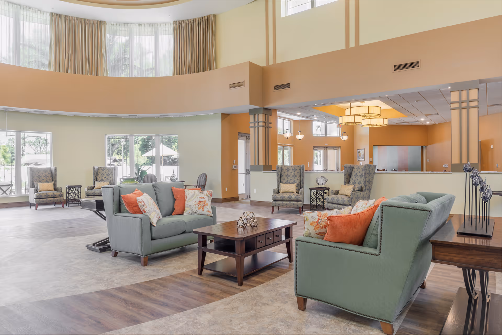 A spacious and well-lit living room area in a senior living facility with large windows, green sofas adorned with orange and floral pillows, patterned armchairs, wooden coffee tables, and modern light fixtures. The room features a mix of wood and tile flooring and soft yellow and beige walls.