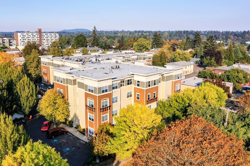 Aerial view of a multi-story senior living facility building surrounded by trees with autumn foliage, parked cars, and a residential neighborhood in the background under a clear sky.