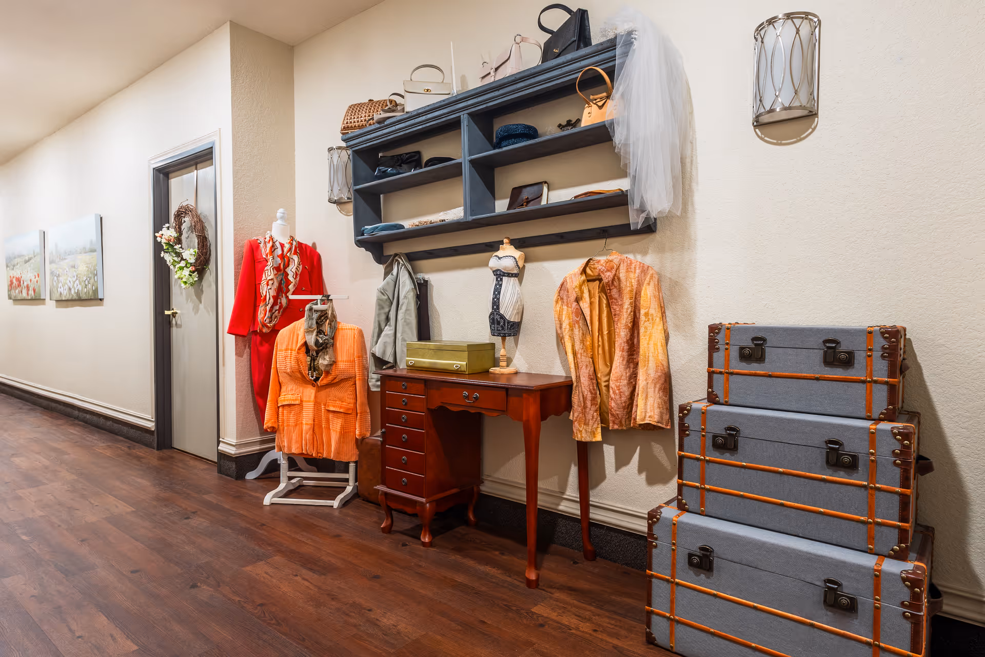 A hallway with wooden flooring and beige walls featuring a display of vintage clothing on mannequins and hangers, a small wooden desk, a wall-mounted shelf with handbags and accessories, and a stack of three vintage-style suitcases. There are also framed floral paintings and a wreath on the wall near a closed door.
