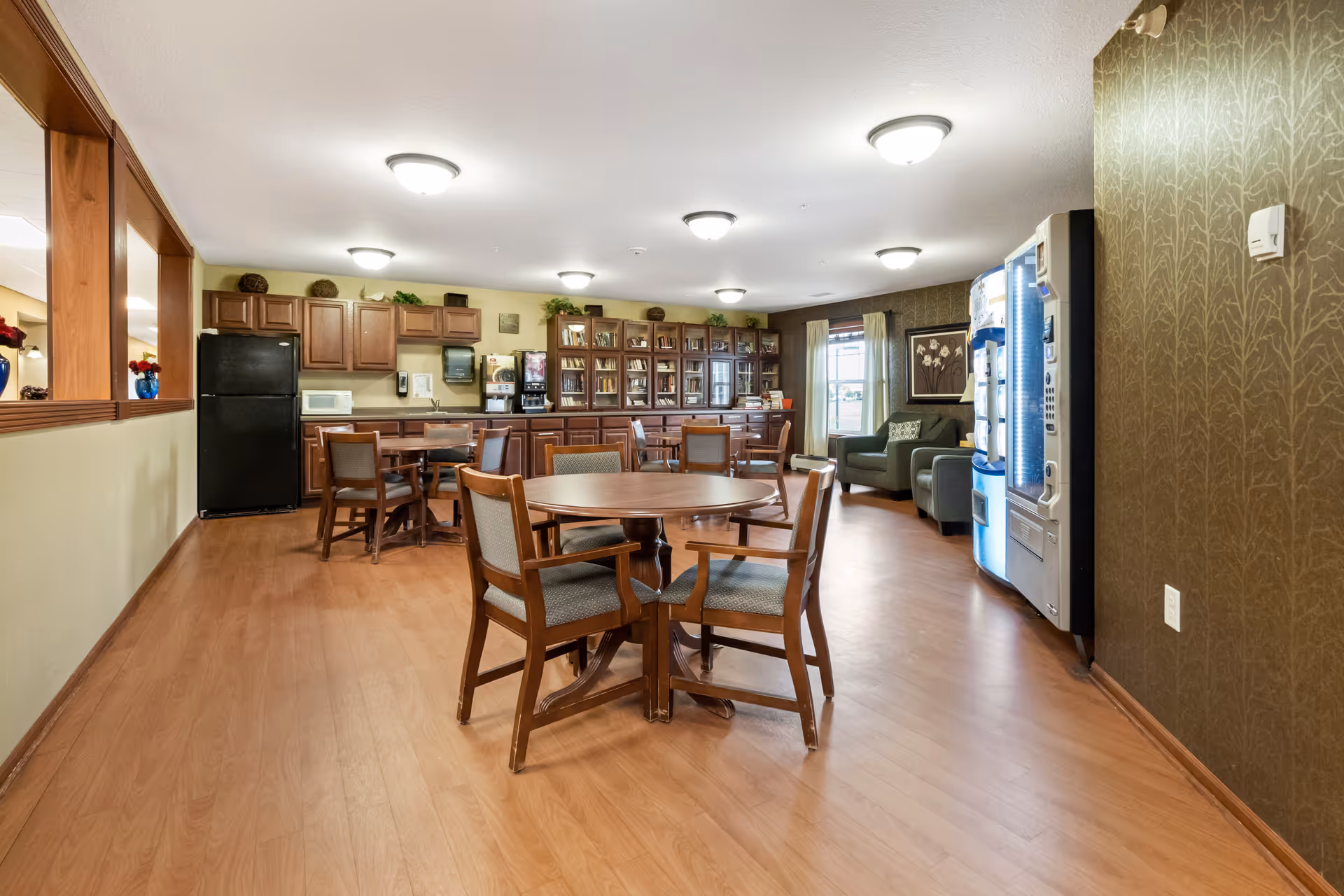 A spacious common area in a senior living facility featuring wooden floors, round tables with chairs, a kitchenette with a black refrigerator, microwave, coffee machine, and cabinets. There is a vending machine on the right side and a seating area with armchairs near a window with curtains. The walls have a patterned wallpaper and the room is well-lit with ceiling lights.