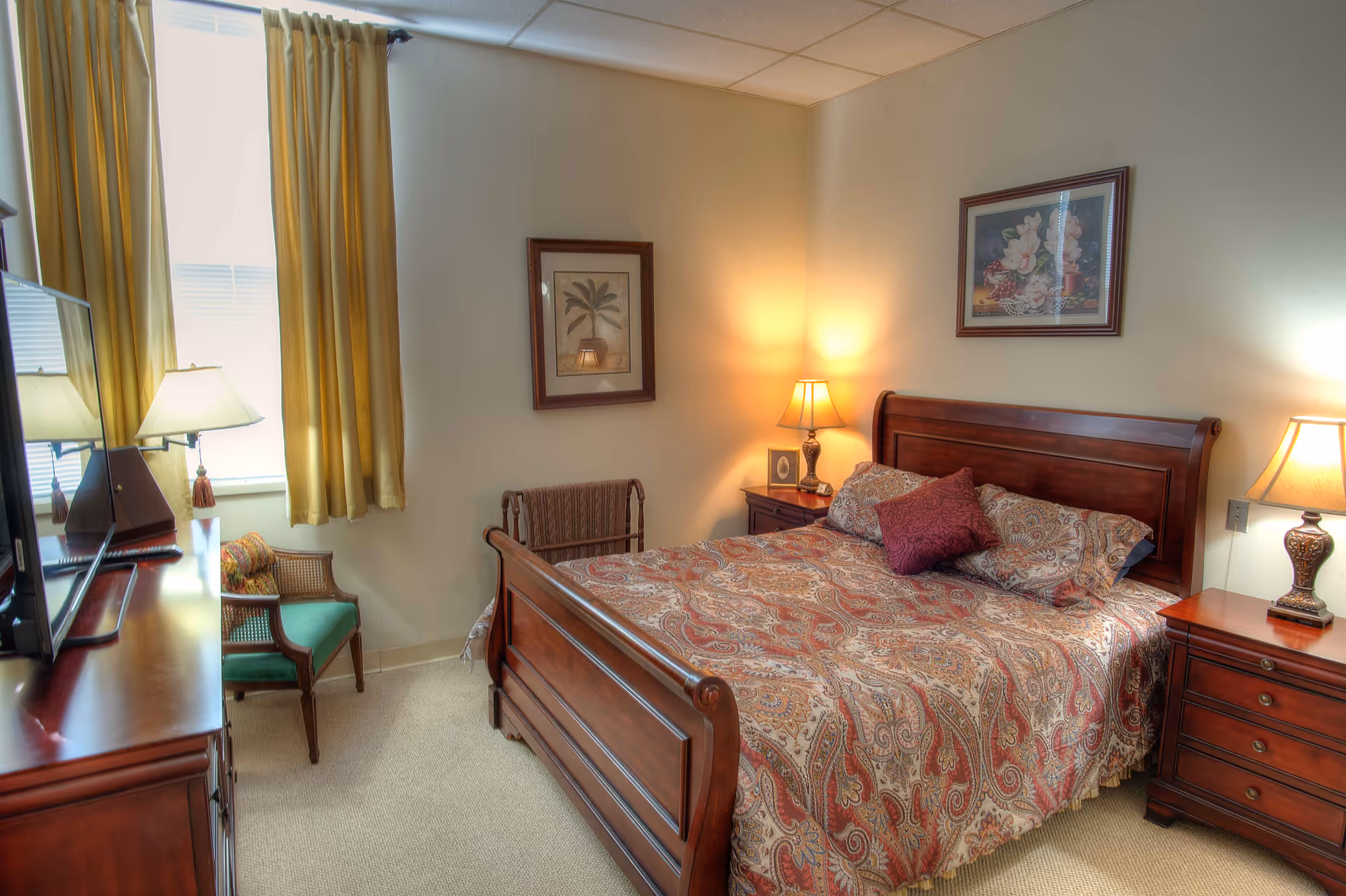 A cozy bedroom with a wooden bed frame and a paisley-patterned bedspread. The room has two wooden nightstands with lamps, a wooden dresser with a TV on top, a green cushioned chair, and two framed pictures on the walls. Yellow curtains cover the window, and the room is softly lit.
