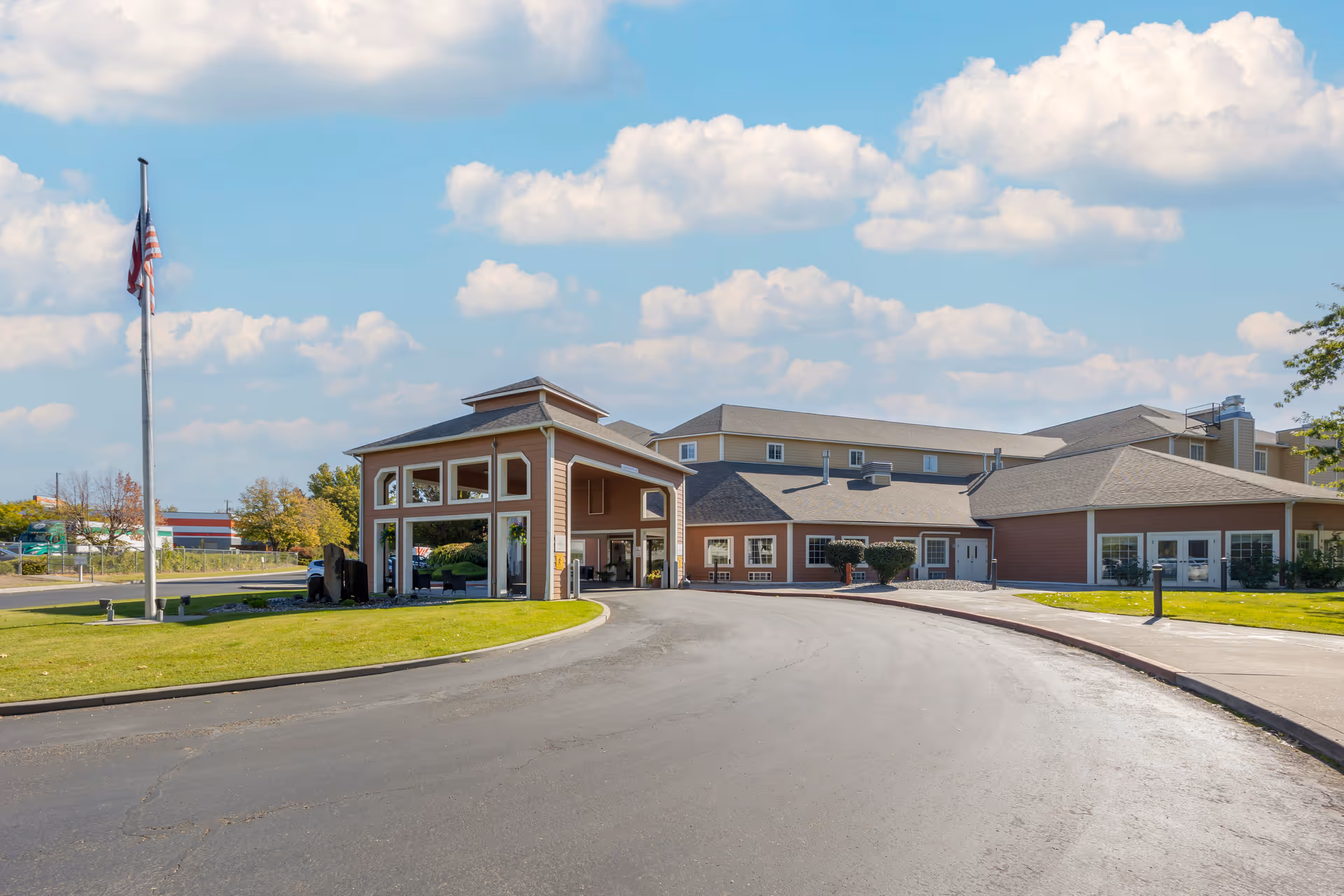 Exterior view of Brookdale Richland senior living facility showing the main entrance with a covered driveway, an American flag on a flagpole, and a clear blue sky with scattered clouds.