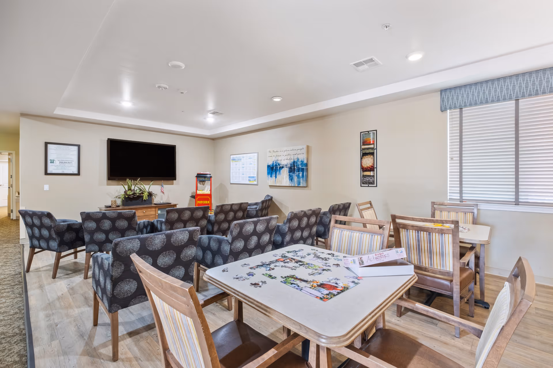 A bright communal lounge with a wall-mounted TV, multiple patterned armchairs facing it, dining tables with chairs and a jigsaw puzzle, and a popcorn machine.