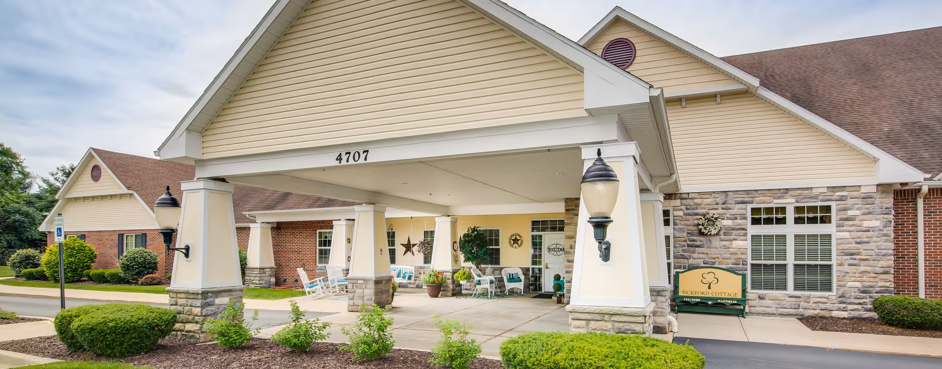 Entrance of Bickford Cottage senior living facility showing a covered drop-off area with stone and brick exterior walls, outdoor seating with chairs and potted plants, and a sign reading 'Bickford Cottage'.