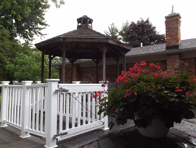 Outdoor view of a wooden gazebo with a shingled roof situated on a deck. The deck has white railings and a gate. In the foreground, there is a large potted plant with red flowers. Behind the gazebo, there is a brick building with a chimney and trees surrounding the area.