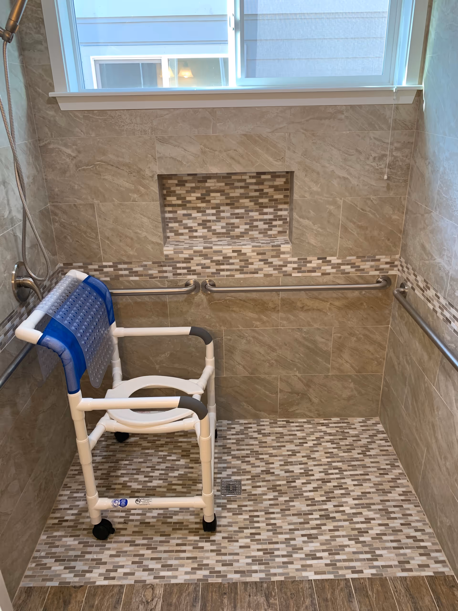Tiled walk-in shower with a mosaic niche, grab bars and a rolling shower chair beneath a window.