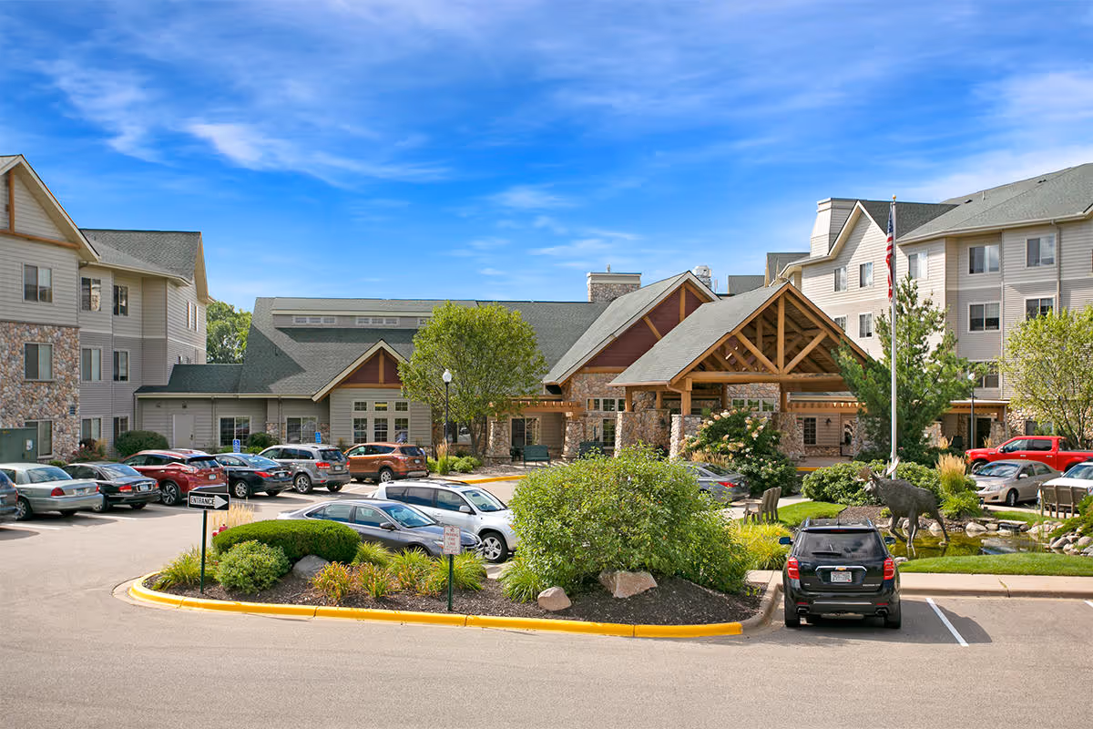 Front exterior of the Timber Hills senior living facility with a lodge-style entrance, parked cars, and landscaped grounds under a blue sky.