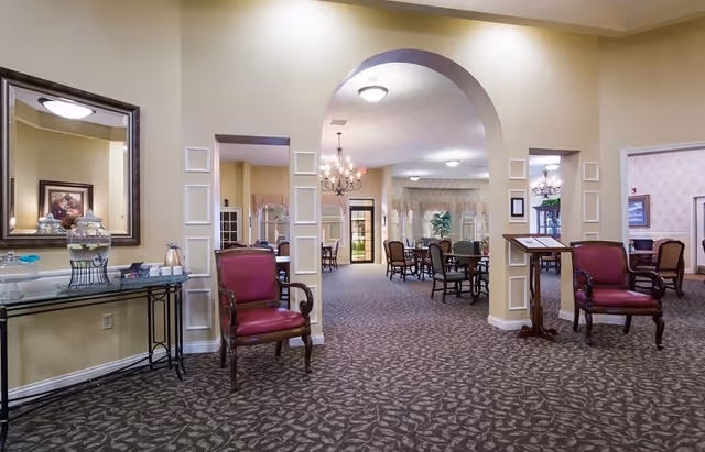 Spacious common dining room with an arched entry, chandeliers, round tables, and upholstered chairs.