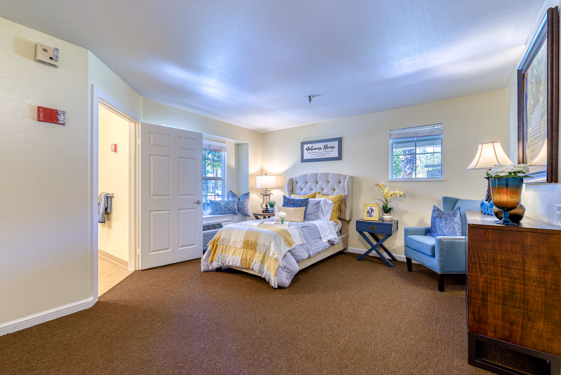 A cozy bedroom in a senior living facility with a single bed featuring a tufted headboard, yellow and gray bedding, and multiple pillows. There is a small blue side table with a plant and a framed photo next to the bed. A blue armchair with a patterned pillow is placed near a wooden dresser with a lamp and decorative items. Two windows allow natural light into the room, and an open door leads to a bathroom. The walls are painted light yellow and the carpet is brown.