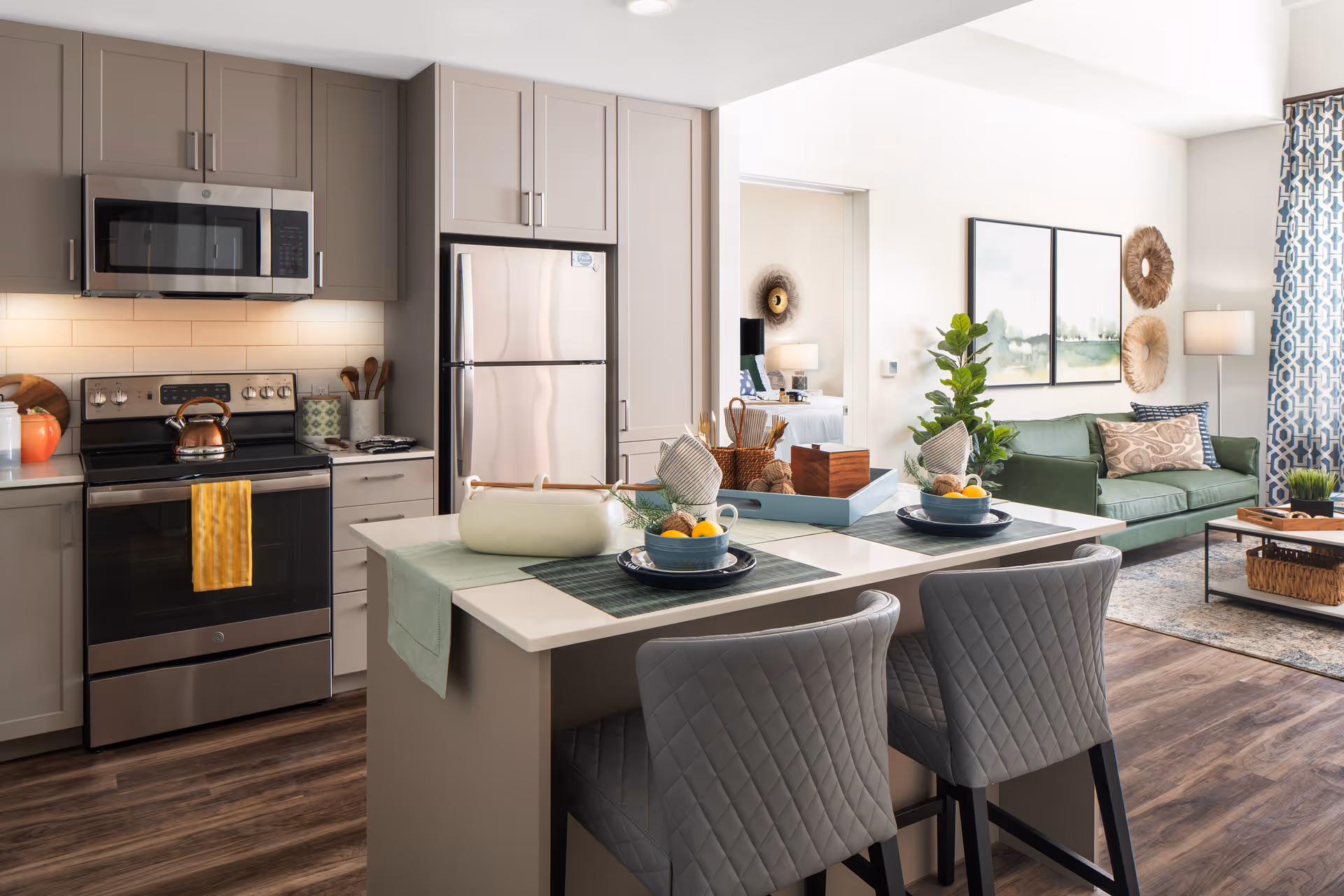 Modern senior living facility interior featuring a kitchen with stainless steel appliances including a microwave, stove, and refrigerator. The kitchen island has two gray quilted chairs and is set with placemats, bowls of fruit, and decorative items. In the background, there is a living area with a green sofa, coffee table, floor lamp, and wall art, with a glimpse into a bedroom.