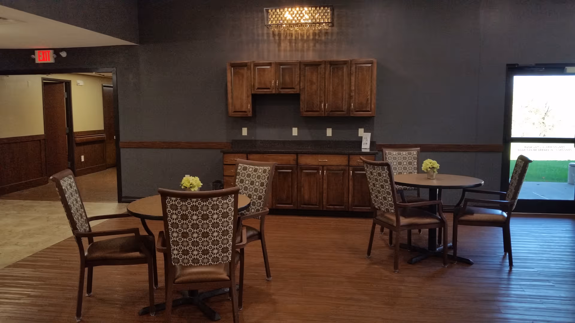 Interior view of a common area with two round tables, each surrounded by four patterned chairs. There are wooden cabinets mounted on a dark gray wall and a small chandelier above. A glass door on the right side leads outside, and a hallway is visible on the left.