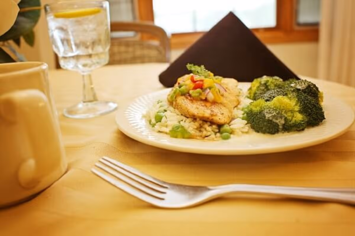 A plate of food on a table with a serving of rice topped with grilled chicken and vegetables, and a side of steamed broccoli. There is a glass of water with a lemon slice, a beige mug, a fork, and a folded dark napkin in the background.