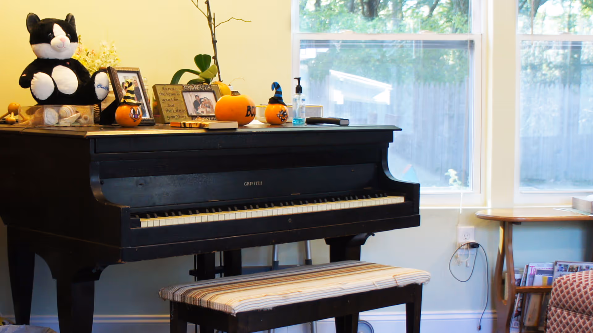 A black upright piano with a striped cushioned bench in front of it. On top of the piano are various decorations including a black and white stuffed cat, small pumpkins, framed photos, a plant, and a hand sanitizer bottle. Behind the piano are large windows showing an outdoor view with trees and a wooden fence. To the right, there is a small wooden table with magazines underneath and part of a patterned armchair is visible.