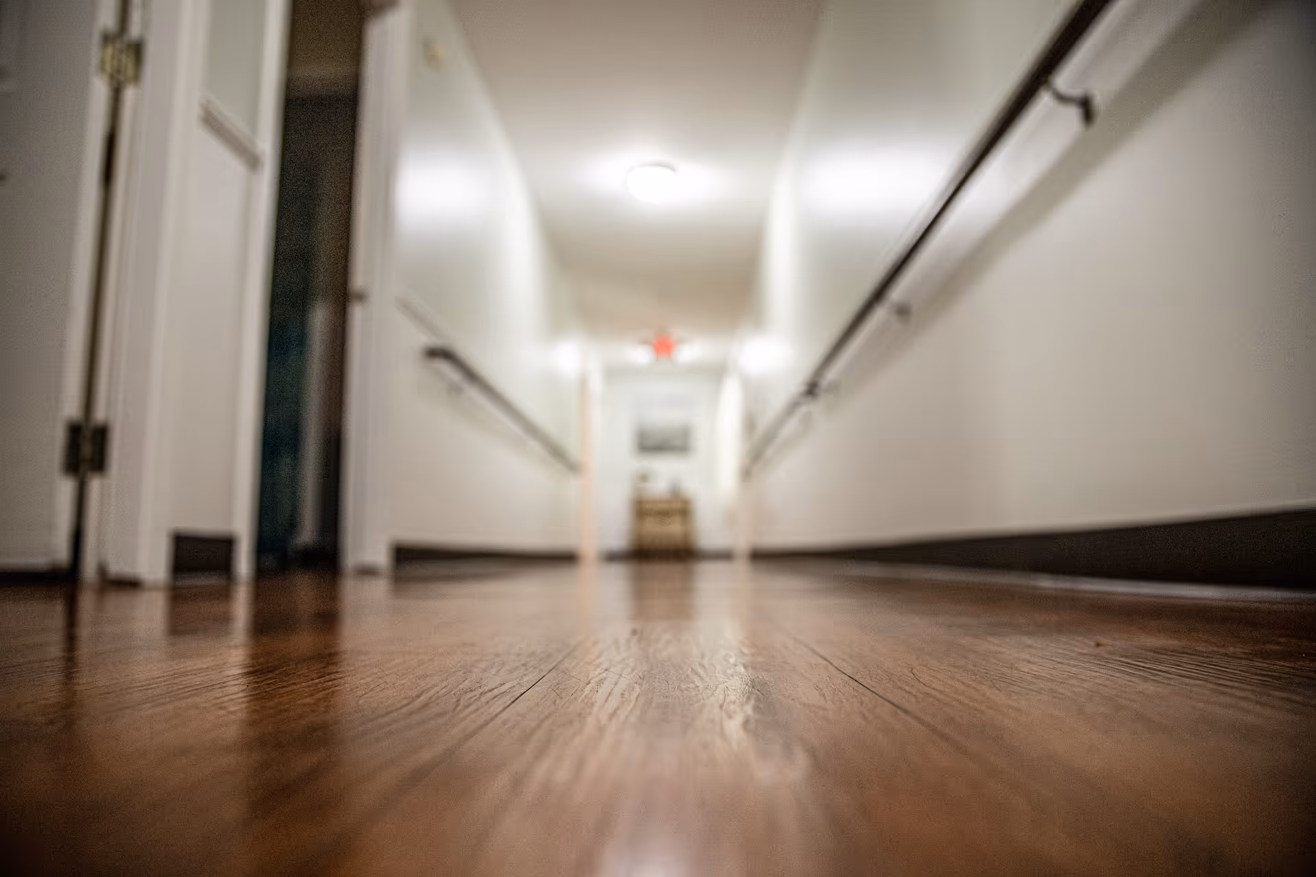 Low-angle view of a long indoor hallway with wooden flooring, white walls, handrails on both sides, and a ceiling light. Doors are visible on the left side and a piece of furniture with a framed picture is seen at the far end of the corridor.