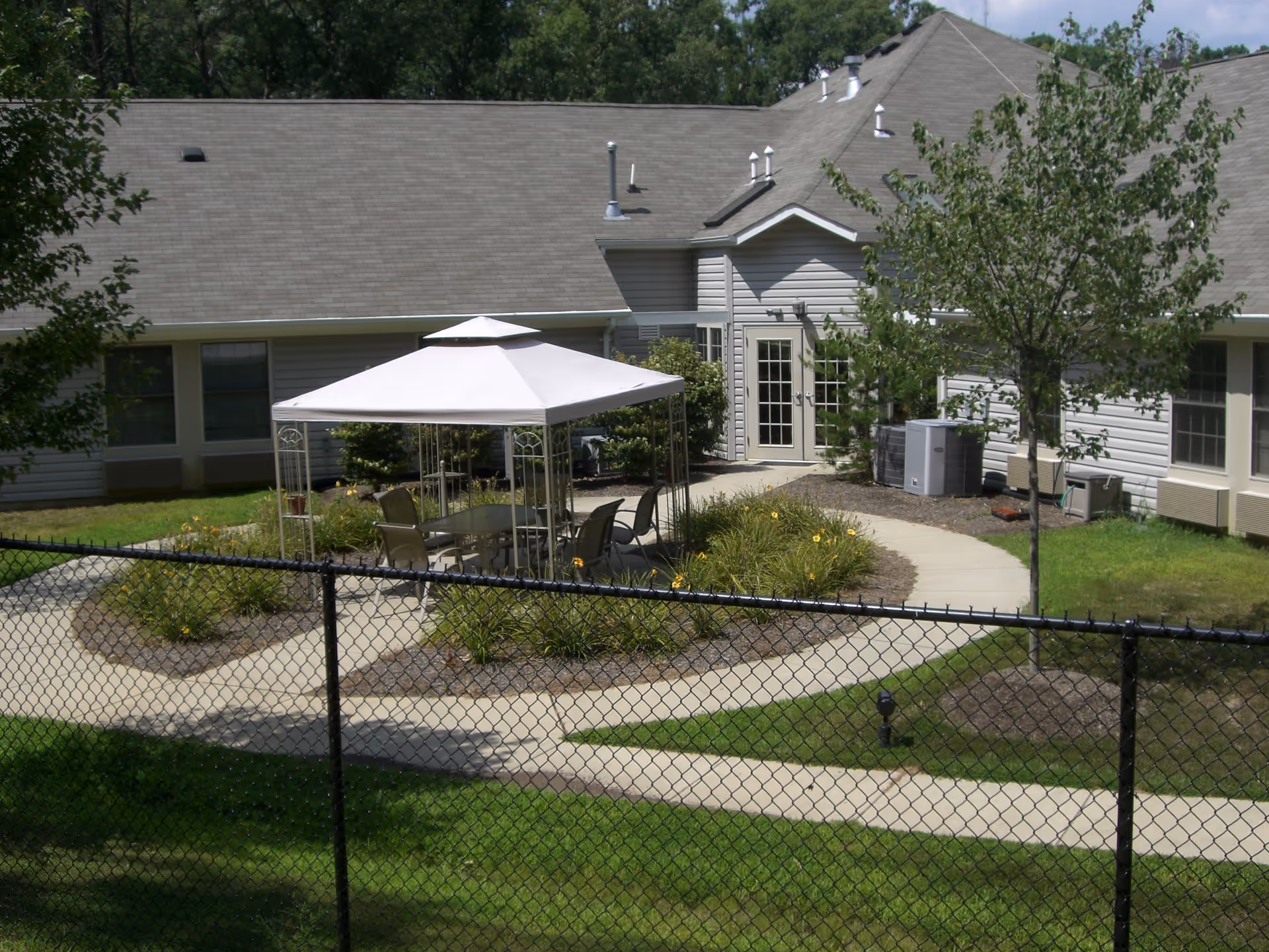 Outdoor patio area at Fenwick Landing Senior Care Community featuring a white canopy over a glass table with several chairs, surrounded by landscaped garden beds and a winding concrete pathway. The building exterior with beige siding and multiple windows is visible in the background, along with some trees and a black chain-link fence in the foreground.