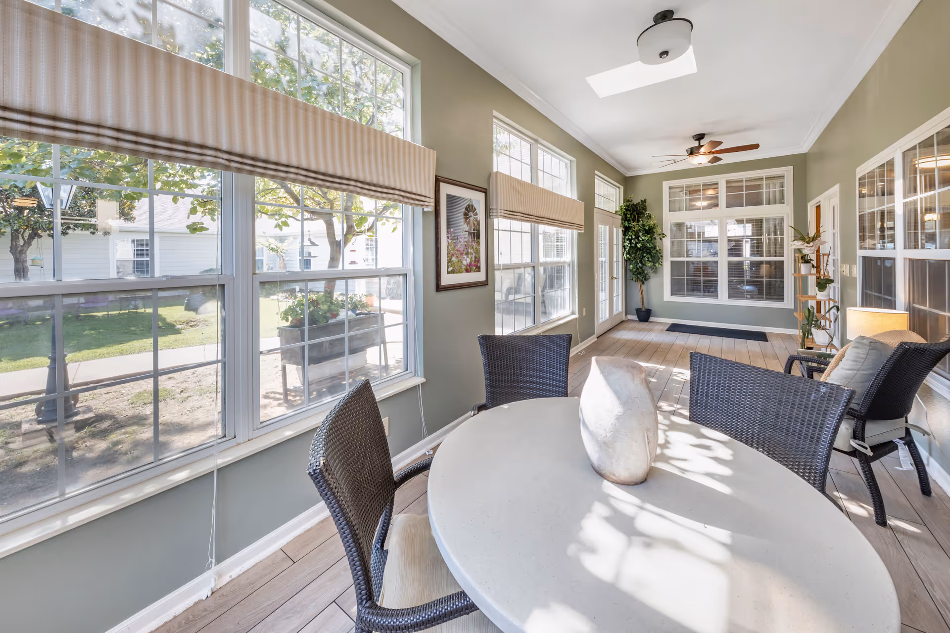 A bright sunroom with large windows showing an outdoor garden area. The room features a round table with four wicker chairs, a decorative vase on the table, a ceiling fan, potted plants, and framed artwork on the wall. The walls are painted light green and the floor is wood.