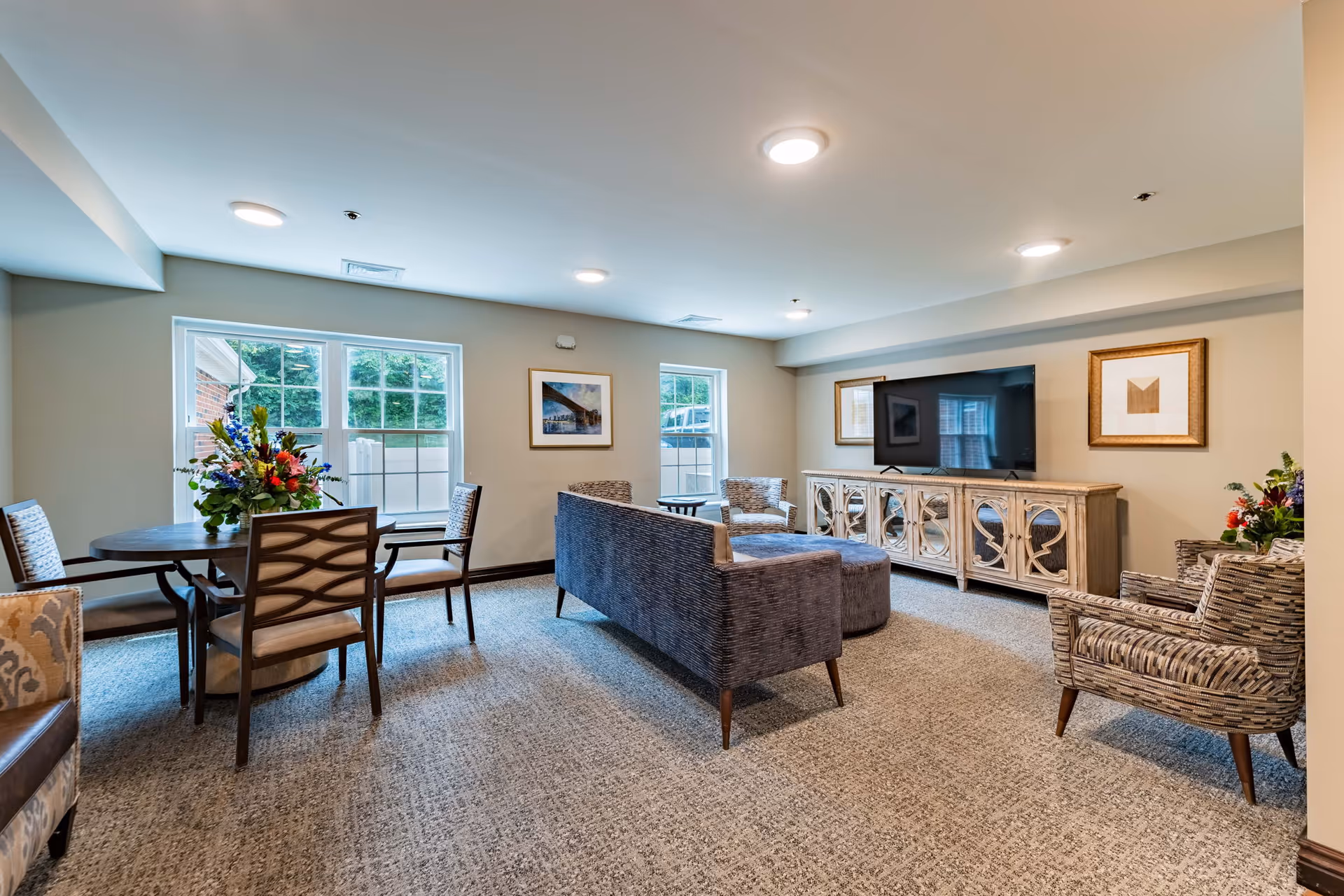 A bright and spacious living room in a senior living facility featuring a large flat-screen TV mounted on a decorative wooden cabinet. The room has a comfortable sofa, two patterned armchairs, a round table with four chairs, and two large windows letting in natural light. The walls are adorned with framed artwork, and there are floral arrangements on the table and beside one armchair.