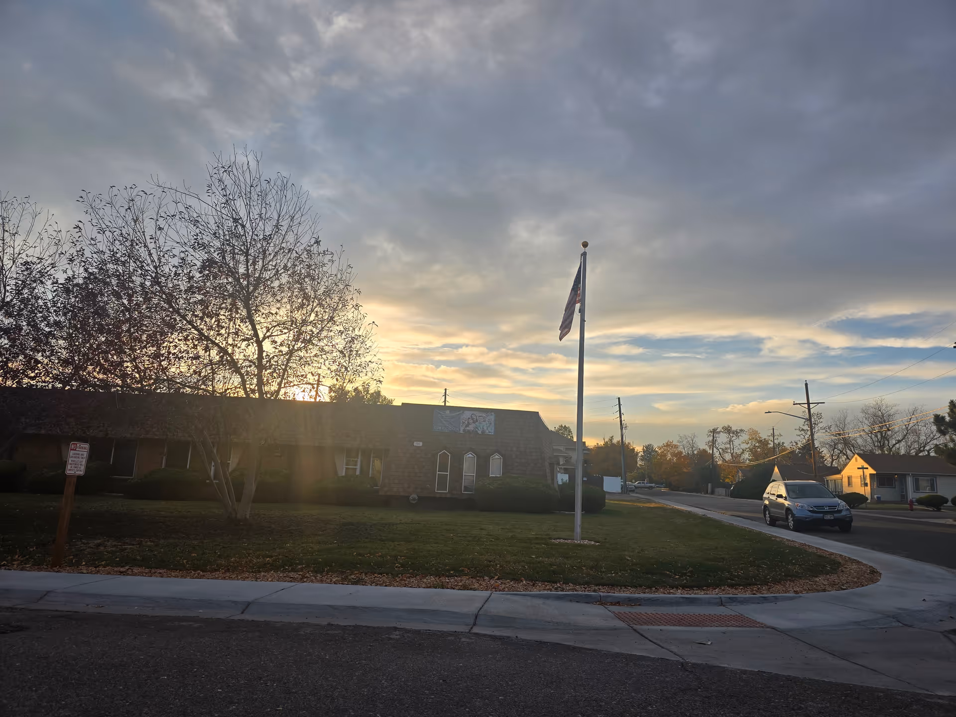 Exterior view of Clear Creek Care Center at sunset with a flagpole displaying the American flag in the front lawn, a tree with sparse leaves, a parked car on the street, and a partly cloudy sky.
