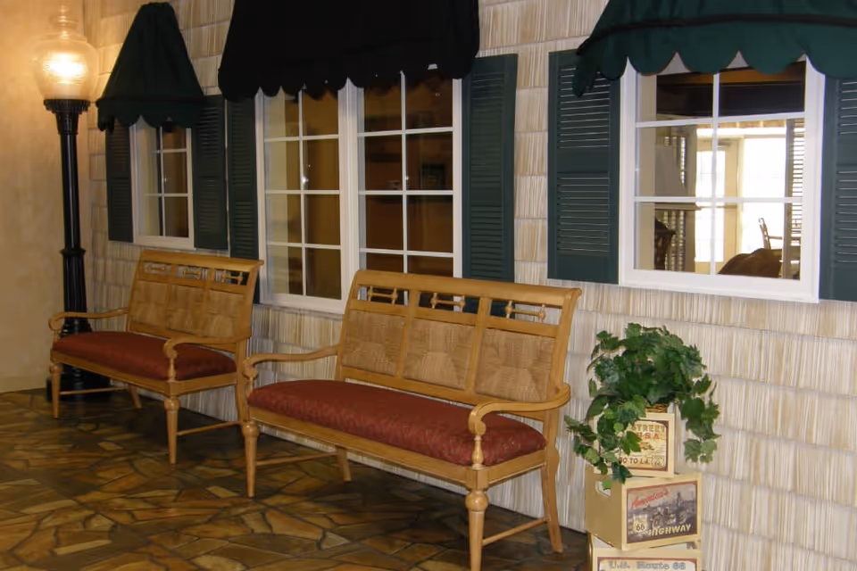 Indoor seating area with two wooden benches featuring red cushions placed against a wall with three windows that have green shutters and green awnings. A tall black lamp with a round light is on the left side, and a decorative plant sits on stacked vintage-style boxes on the right side.