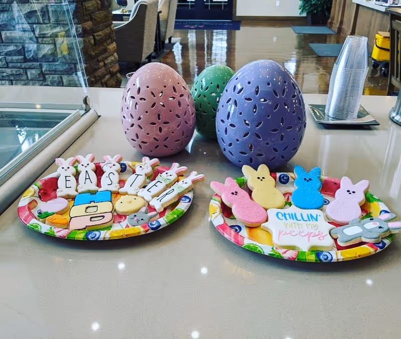 Two colorful plates with Easter-themed cookies including bunny-shaped cookies spelling out 'EASTER' and marshmallow peeps, displayed on a countertop with three decorative perforated egg-shaped ornaments in pink, green, and purple in the background. A stack of clear plastic cups is also visible on the right side of the counter.