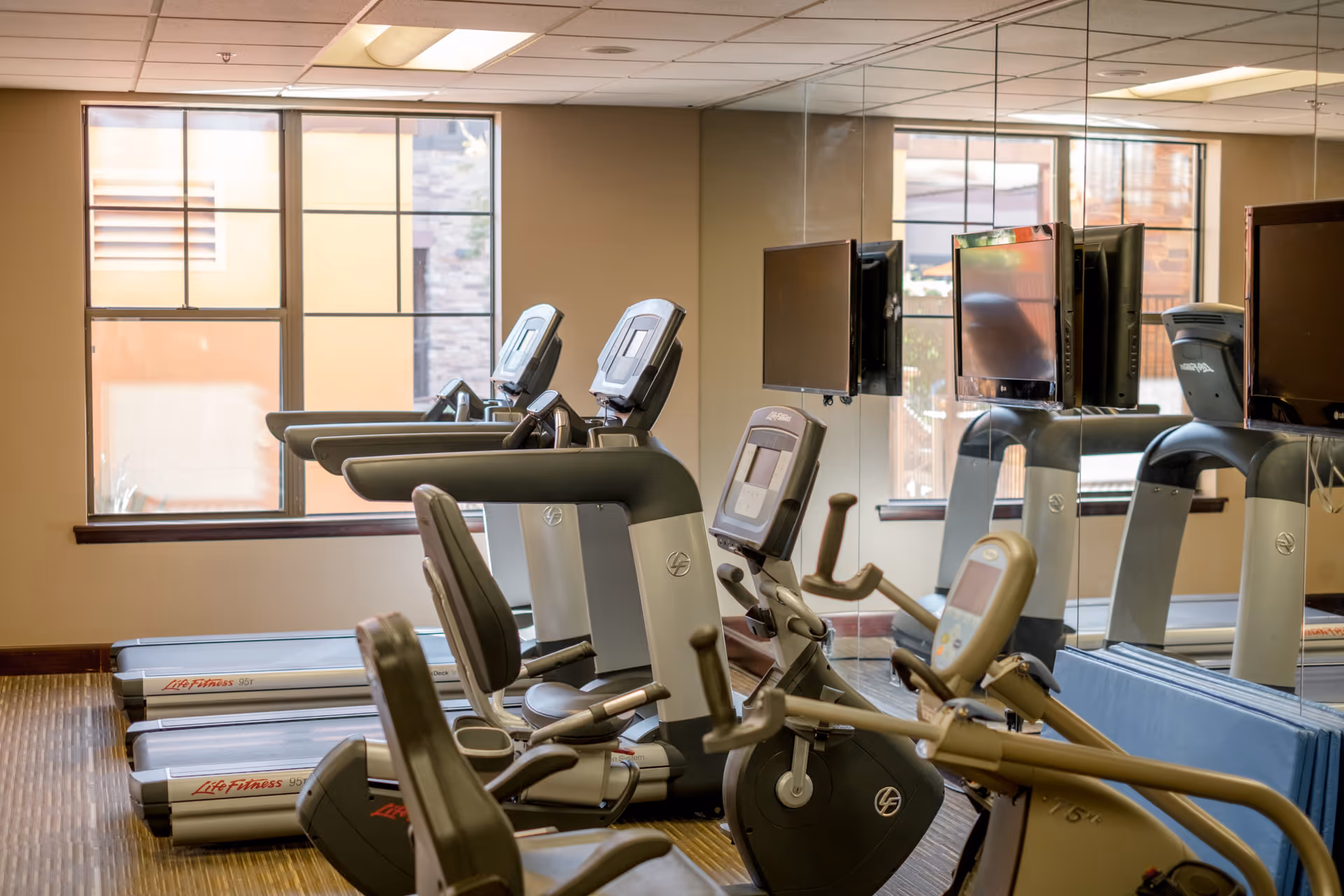 A fitness room with several exercise machines including treadmills and stationary bikes, large windows letting in natural light, and a wall covered with mirrors reflecting the equipment.