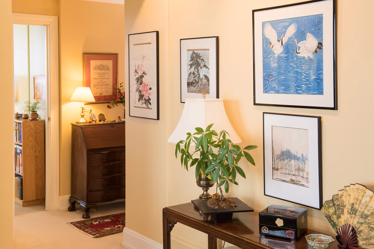 A warmly lit interior hallway corner featuring a wooden console table with a potted plant, a table lamp, a decorative box, and a decorative fan. The walls are adorned with framed artwork including paintings of cranes, flowers, and landscapes. In the background, there is a wooden chest of drawers with a lamp and framed certificate on the wall above it, and a glimpse into another room with bookshelves and plants.