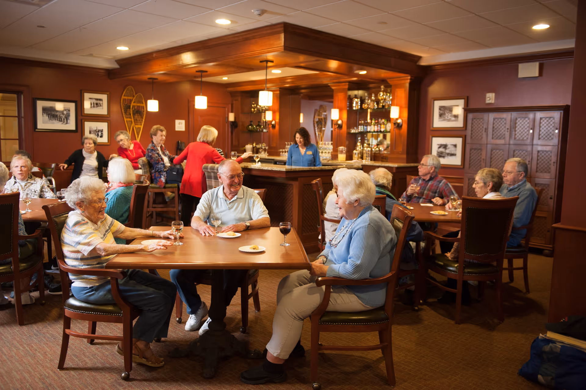 Residents socializing around tables and at a bar in a warm, wood-paneled common dining/lounge area.