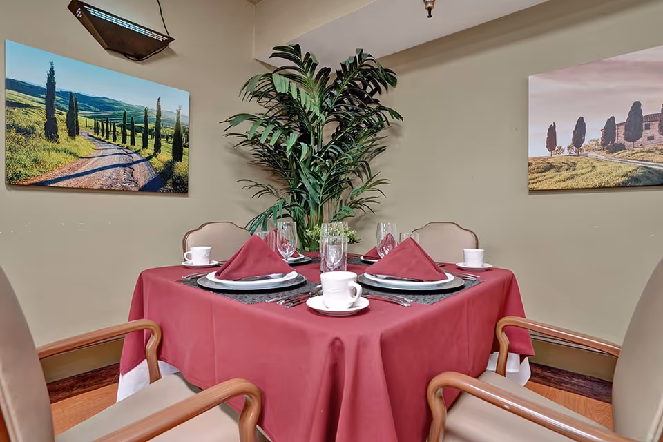 A dining table set for four with a burgundy tablecloth, matching folded napkins, white plates, cups, and glasses. The table is surrounded by beige chairs with wooden armrests. Behind the table is a large green plant and two landscape paintings on the beige walls.