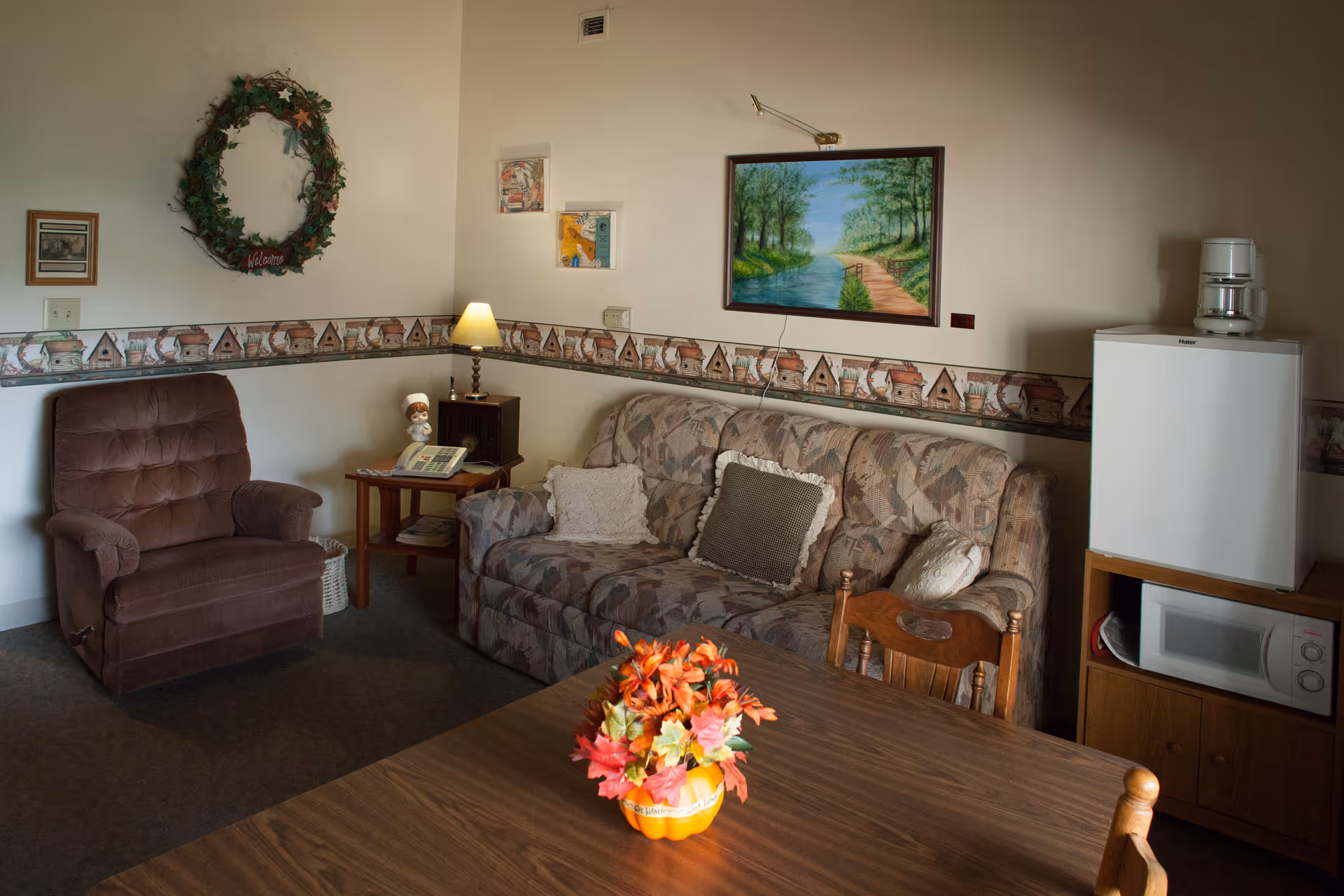 A cozy living room area with a patterned sofa adorned with three pillows, a brown armchair, a wooden side table with a lamp and a telephone, a small refrigerator with a coffee maker on top, and a microwave below. The walls are decorated with a wreath, framed pictures, and a painting of a river and trees. A wooden table with a floral centerpiece is in the foreground.