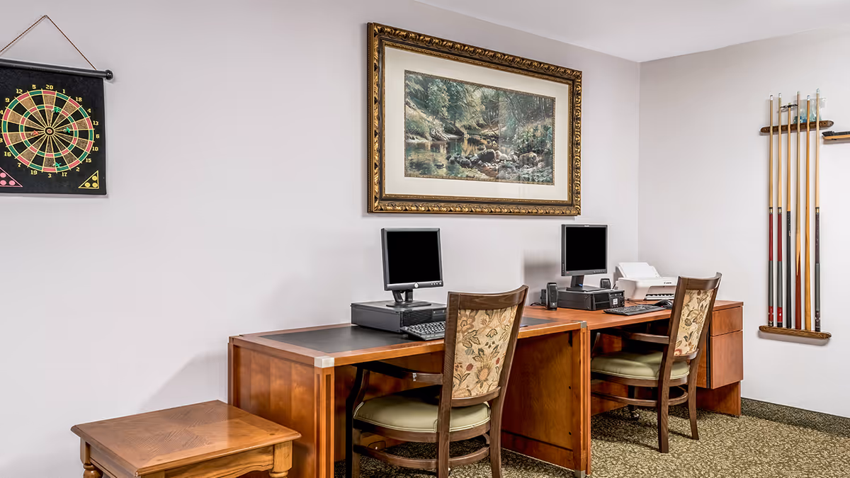 Two wooden computer workstations with chairs, monitors and a printer in a sitting/office area with a dartboard and pool cues on the walls.