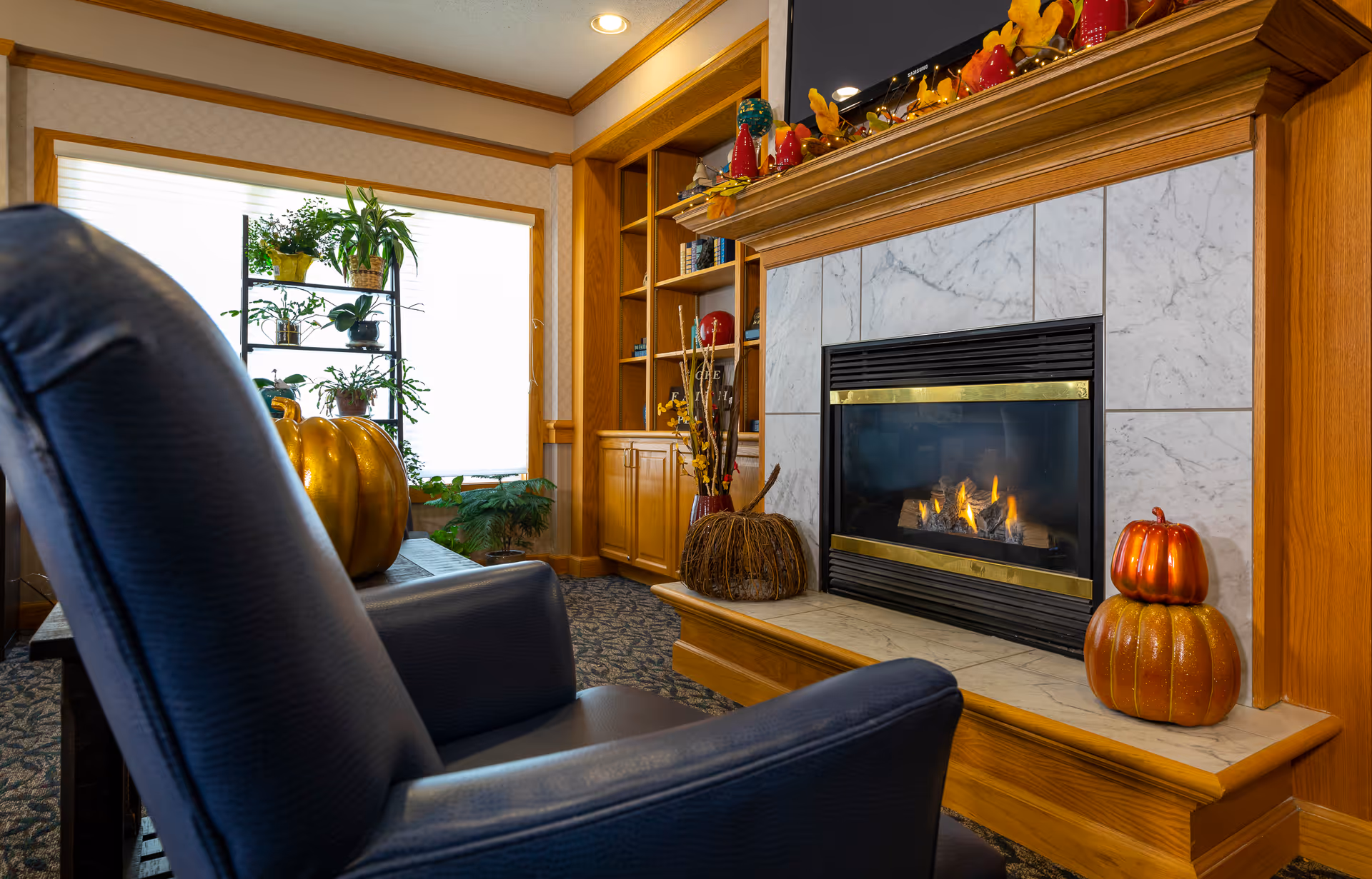 Cozy living room area with a black leather armchair facing a lit fireplace surrounded by marble tiles and wooden mantel decorated with autumn-themed ornaments and pumpkins. A wooden built-in bookshelf and a window with plants on a metal shelf are visible in the background.