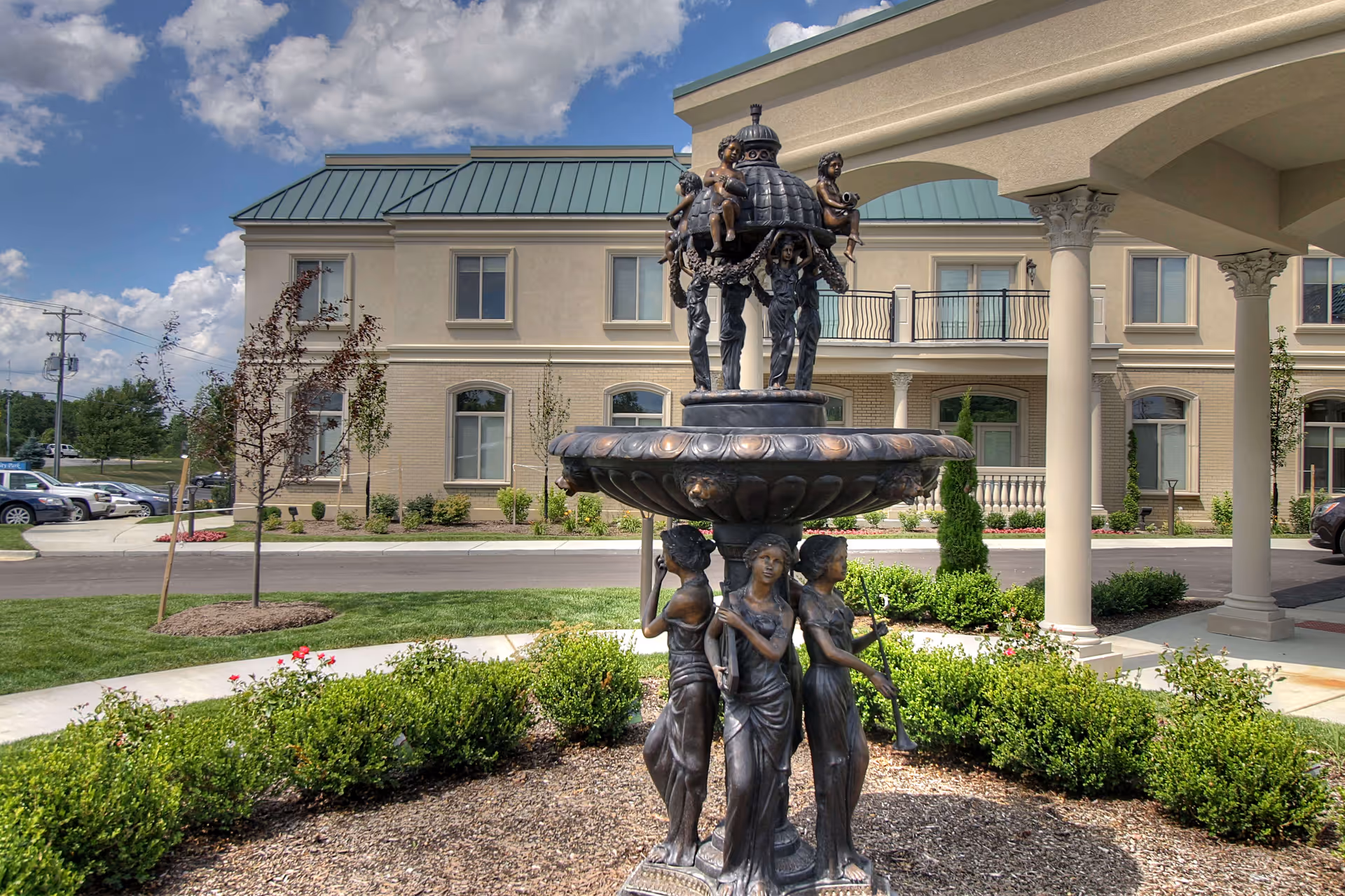 Outdoor view of a senior living facility named Tranquility Estates featuring a decorative bronze fountain with statues of women and children in the foreground, manicured bushes, a tree, and a beige building with green roof and columns in the background under a partly cloudy sky.