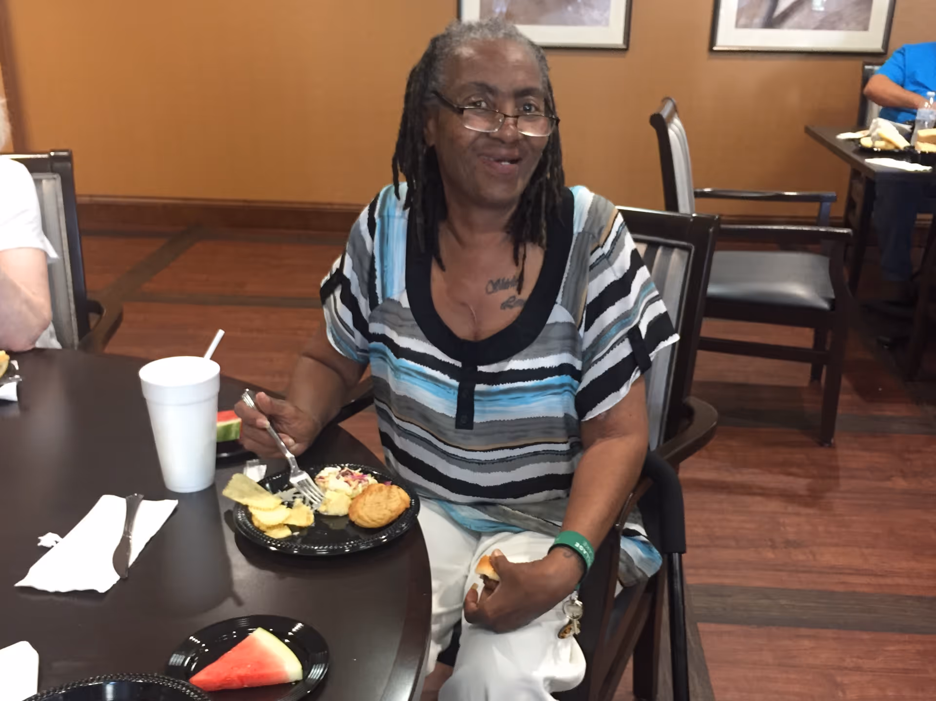 A woman sits at a dining table in a dining room, smiling and eating from a plate of food.
