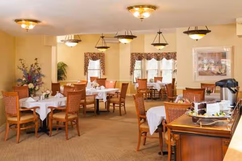 A well-lit dining room with round tables, white tablecloths, wooden chairs, and a side buffet.
