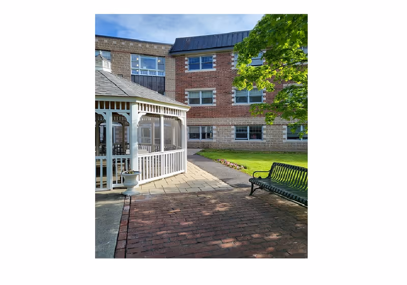 Brick nursing facility courtyard with a white gazebo, a green bench, a tree, and walkways.