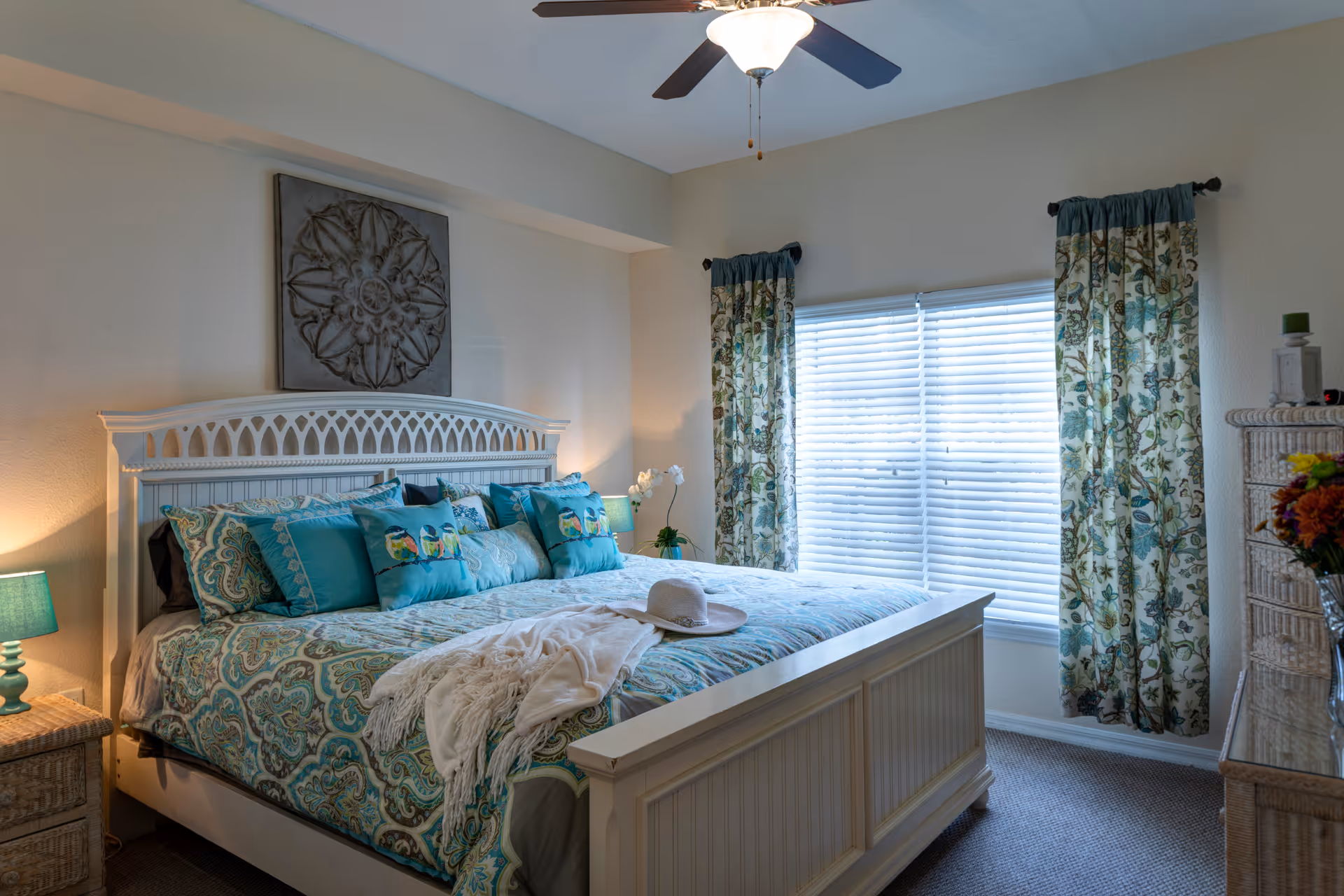 A cozy bedroom with a large white wooden bed adorned with teal and patterned bedding and multiple pillows. A white throw blanket and a hat are placed on the bed. The room has a ceiling fan with a light, two windows with floral curtains, a wicker nightstand with a teal lamp, and a wicker dresser with a vase of flowers.