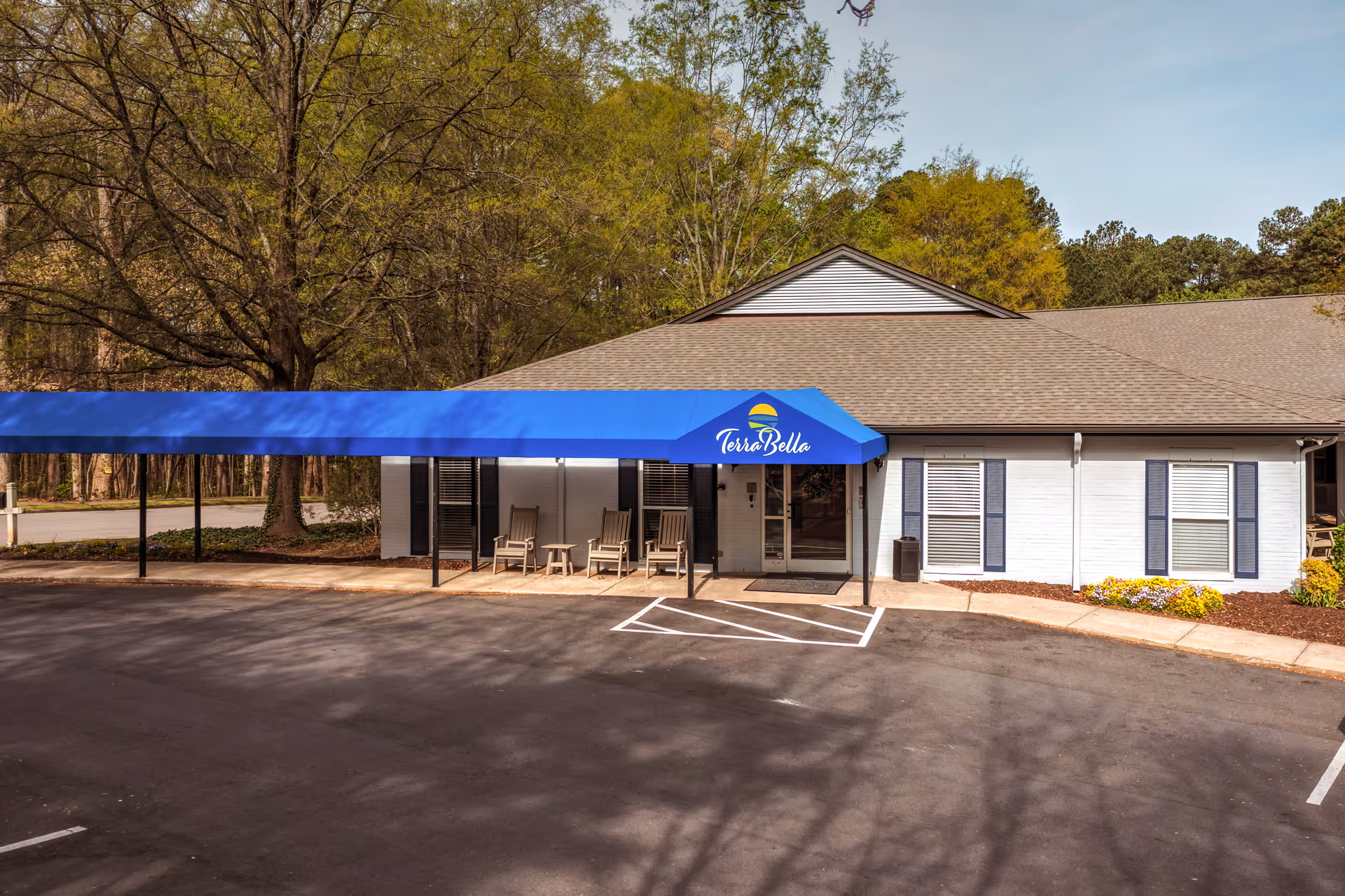 Exterior view of TerraBella Northridge facility showing a single-story building with a blue awning over the entrance. There are several chairs and small tables on the porch area, surrounded by trees and landscaping with flowers. The parking lot is visible in the foreground.