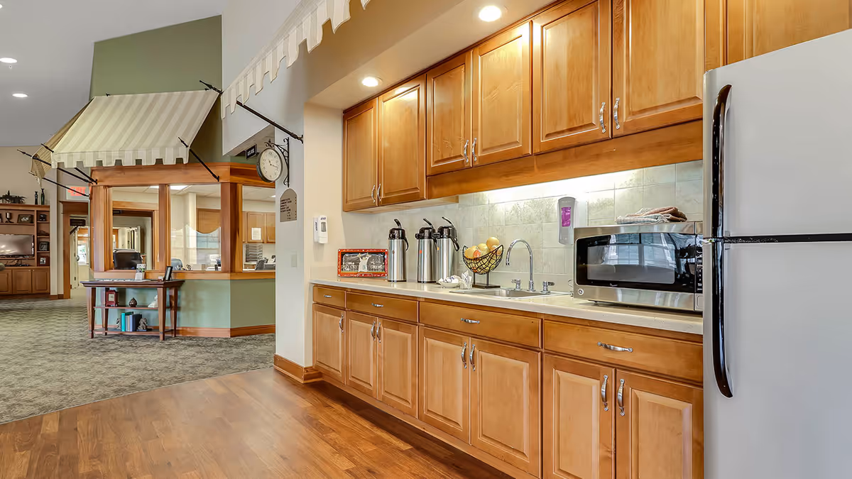Interior view of a kitchenette area in an assisted living facility with wooden cabinets, a countertop with a sink, coffee dispensers, a fruit basket, a microwave, and a refrigerator. In the background, there is a reception or office window with a striped awning above it and a clock on the wall.