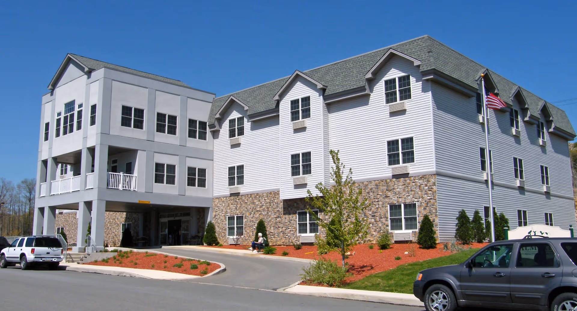 Exterior view of a multi-story senior living facility named The Palmerton with a combination of stone and white siding on the building. There are several windows, a covered entrance, landscaped areas with small trees and shrubs, an American flag on a flagpole, and parked cars in front under a clear blue sky.