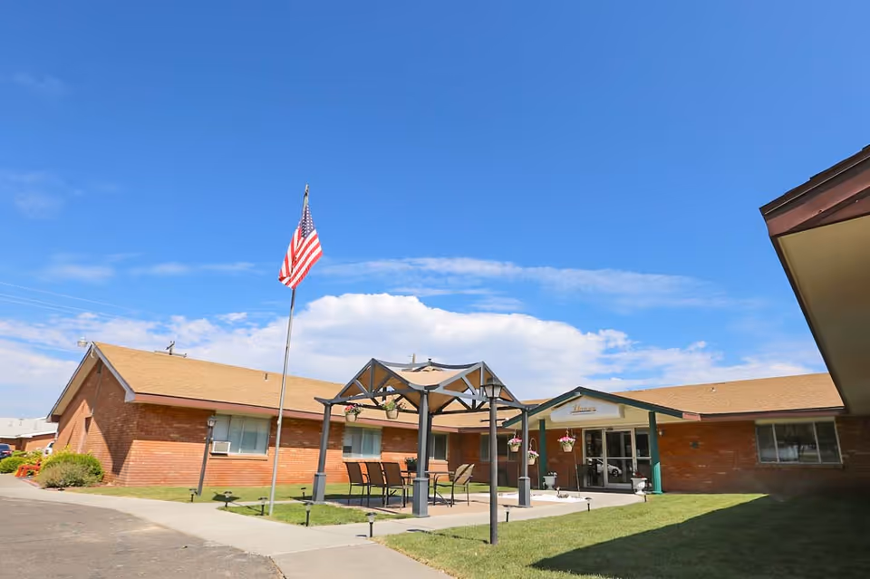 Exterior view of Heritage Assisted Living Of Twin Falls facility showing a single-story brick building with a tan roof, an American flag on a flagpole, a covered outdoor seating area with chairs and hanging flower pots, and a clear blue sky with some clouds.
