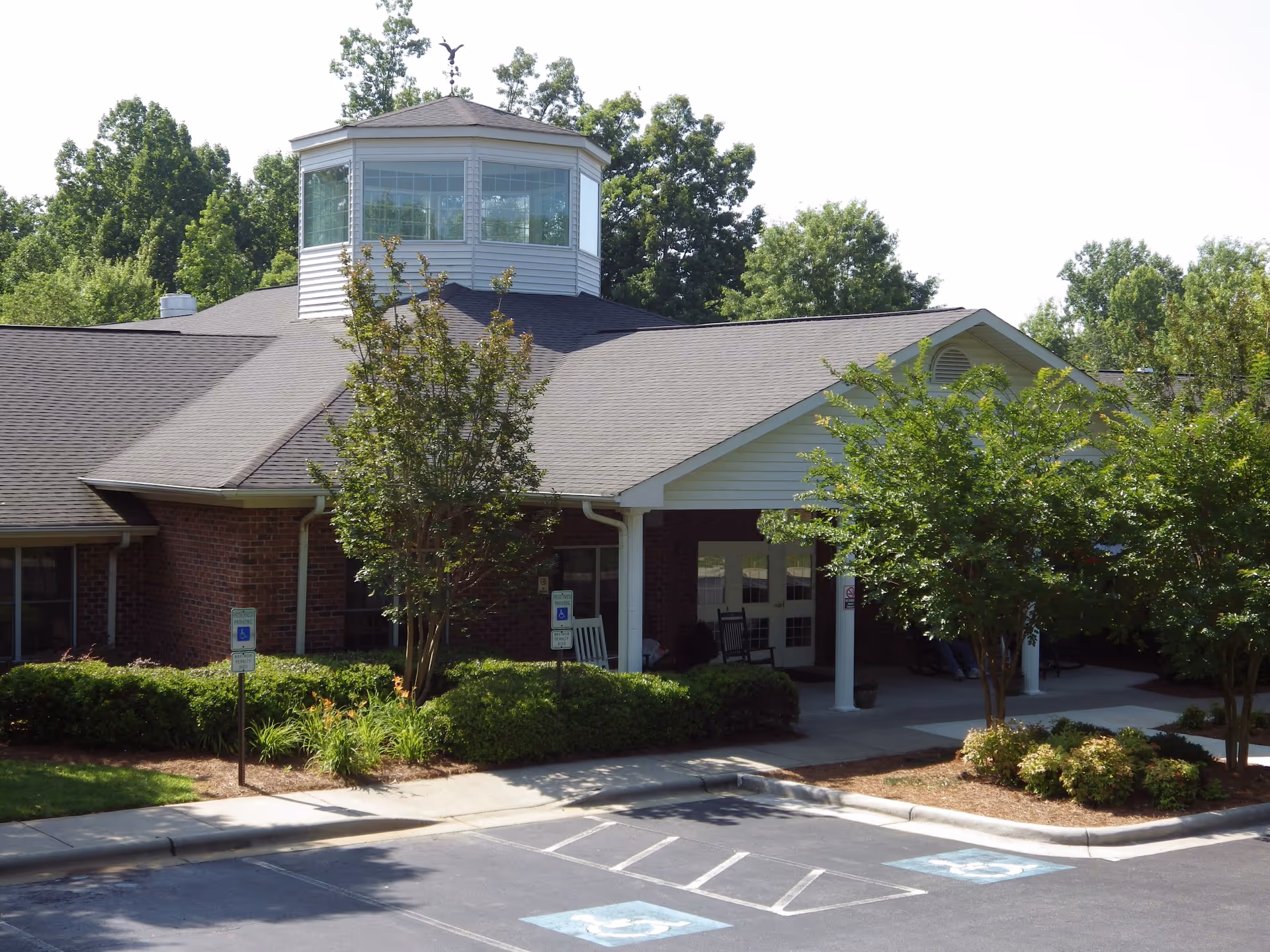 Front entrance of a brick senior living building with a covered entry, rocking chairs, landscaping, and marked wheelchair parking spaces.