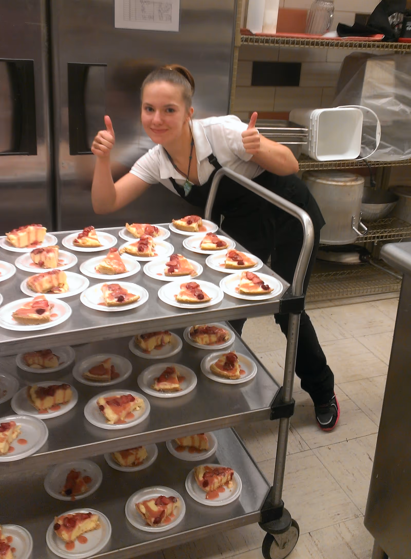A smiling staff member gives two thumbs up next to a cart stacked with many plated dessert slices in a commercial kitchen.
