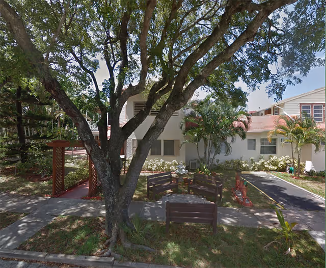 Exterior view of a residential building with a large shade tree, benches around a small landscaped courtyard and paved walkways.