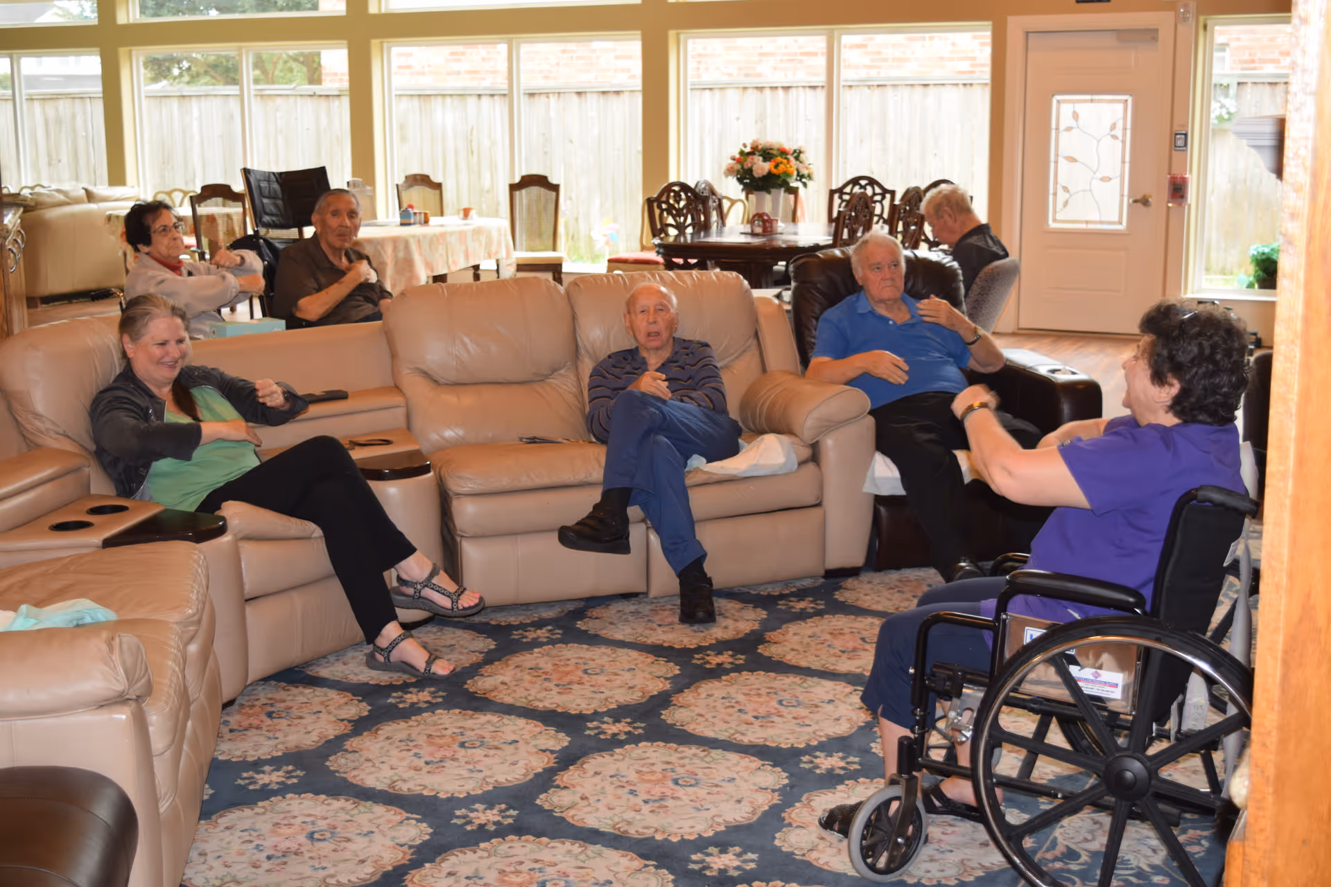A group of elderly people sitting and interacting in a spacious living room with large windows, beige leather sofas, a patterned carpet, and a dining table with chairs in the background. One person is seated in a wheelchair.