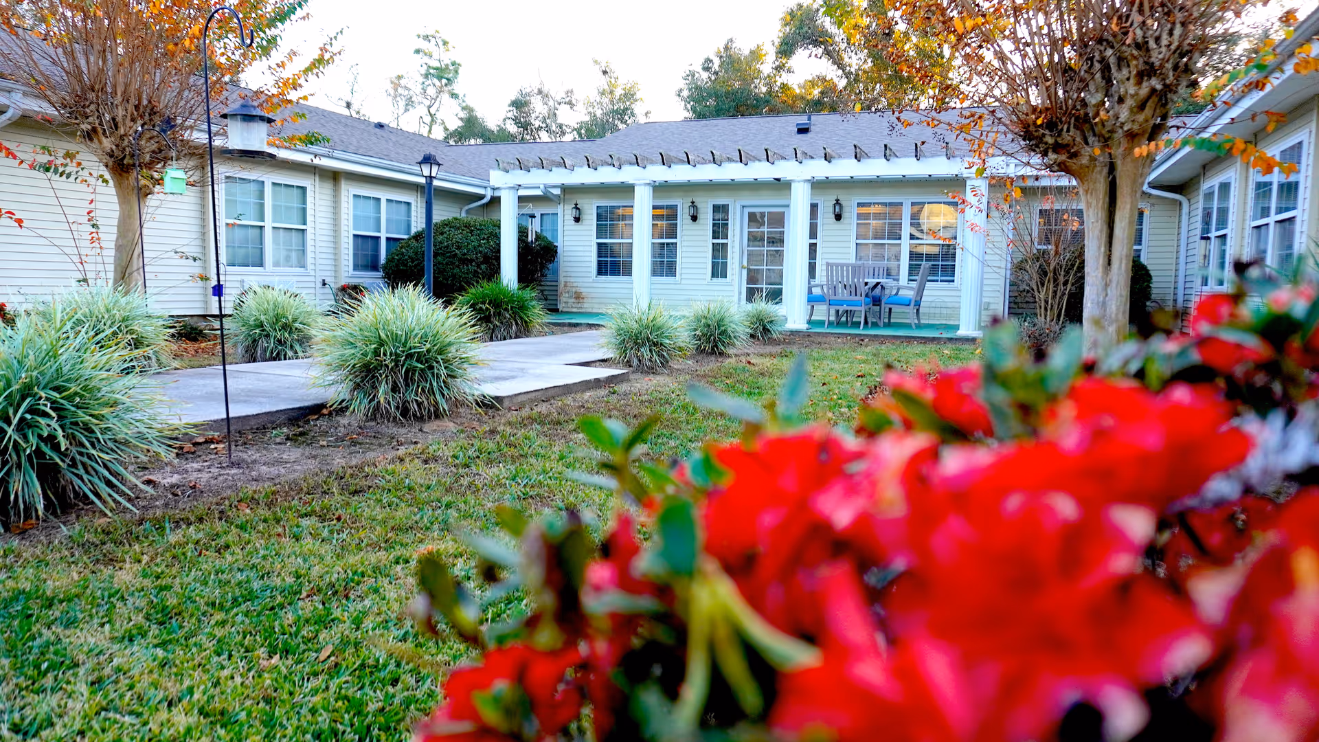 Outdoor view of a single-story senior living facility building with beige siding, white trim, and a covered patio area with outdoor seating. The foreground features green grass, ornamental grasses, and vibrant red flowers, with trees on either side of the walkway leading to the entrance.