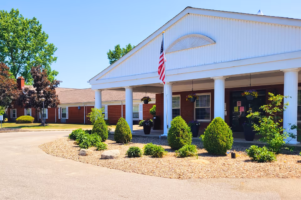 Front exterior view of a single-story brick building with white columns and a covered entrance. There is an American flag on a pole near the entrance, surrounded by neatly trimmed bushes and plants. The sky is clear and blue, and there are trees in the background.